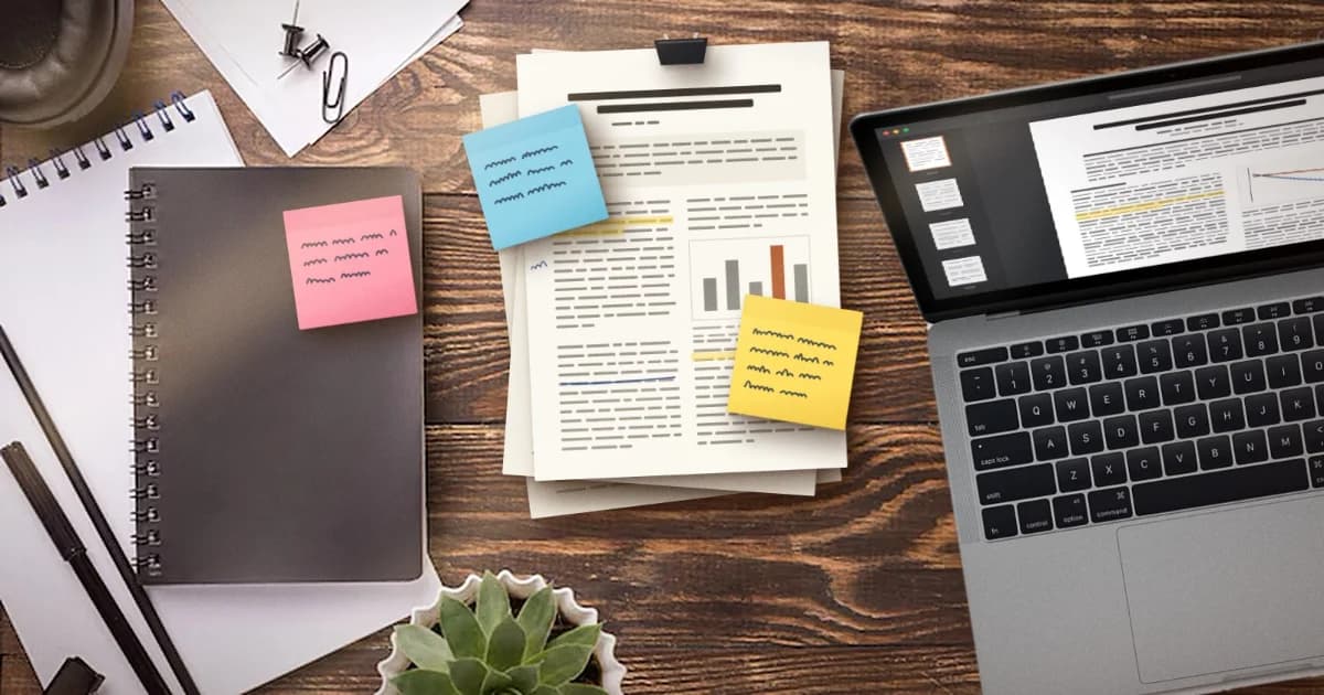 A stack of printed research papers with colored sticky notes beside a laptop